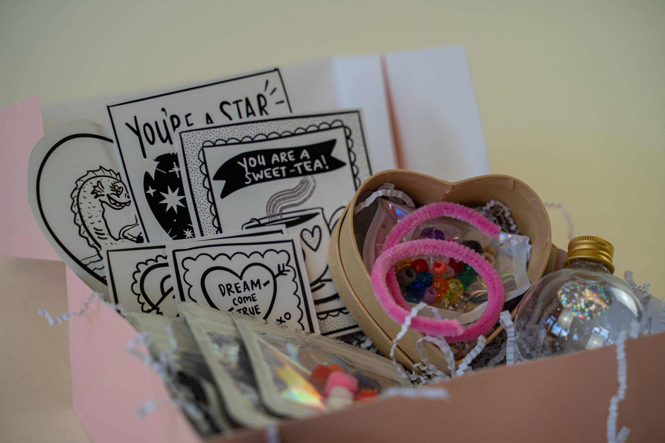 Miss Jasmine Tea's Valentine's Kit comes with a gift box containing greeting cards, a pink heart-shaped bead container, a small bottle with a shiny top, and pink pipe cleaners, all nestled in soft shredded paper.