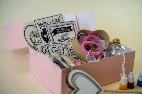 Unbox Miss Jasmine Tea's Valentine's Kit on a beige surface to find a pink box containing decorative cards, a wooden heart, a small bottle of clear liquid, and charming craft items.