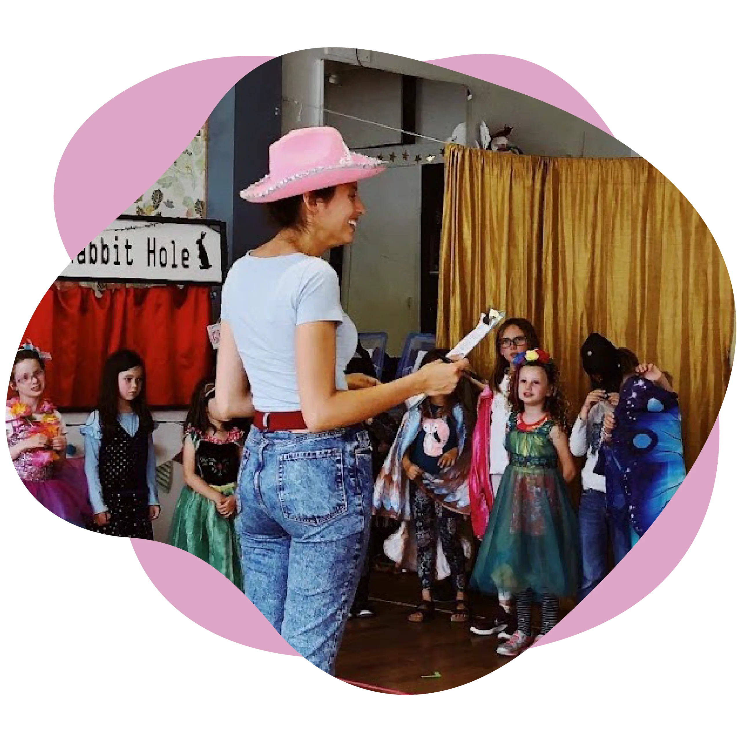 Person in a pink cowboy hat leads a group of children in costumes, inviting them to "Join Our Team." They're inside a room decorated with a "Rabbit Hole" sign and framed by a bright yellow curtain.