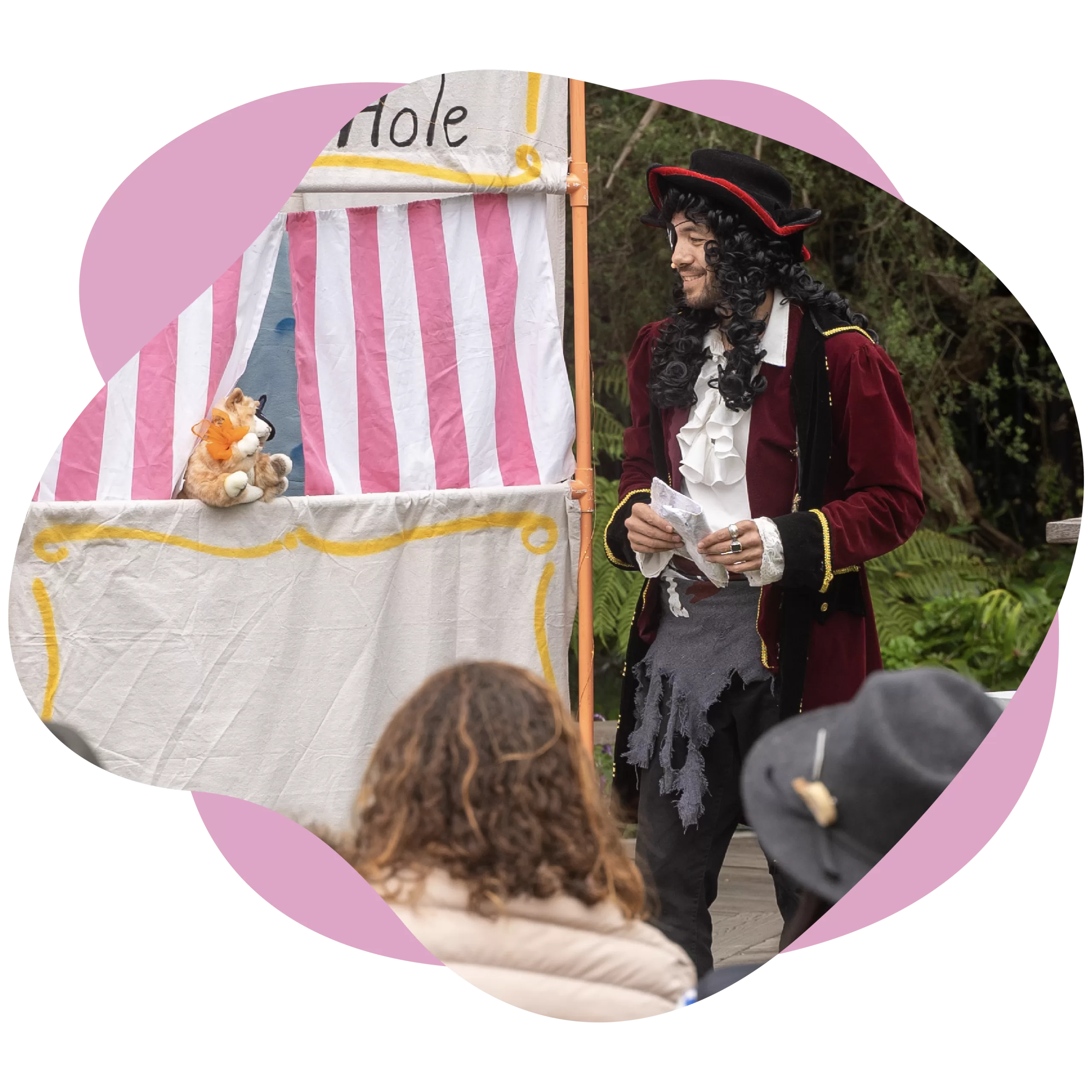 A performer in pirate attire stands near a puppet show stage, holding papers. A toy tiger puppet peeks from the stage as the audience watches, with some people wearing hats.