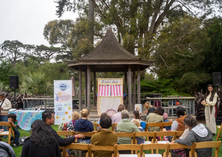 Outdoor puppet show with an audience seated in front of a small stage labeled "Rabbit Hole." A host stands to the right, trees and a gazebo are in the background.