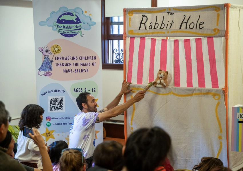 A man holds a puppet on stage at a children's theater event. A banner reads "The Rabbit Hole" with audience members watching.