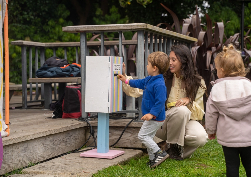 A woman crouches next to two young children at a colorful outdoor box. One child interacts with the box. A wooden deck and greenery are in the background.