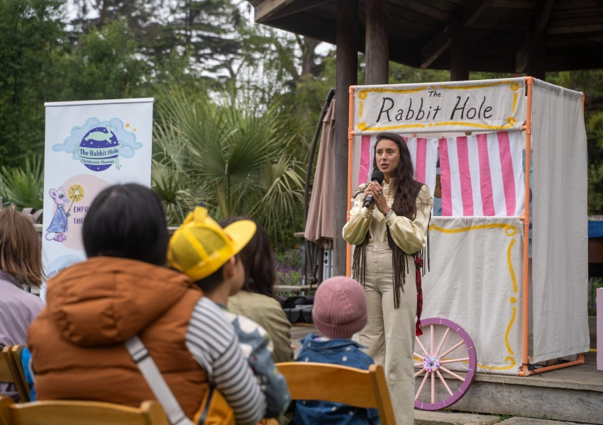 A woman speaks to an audience of children outdoors, standing beside a sign and a small pink and white puppet stage labeled "The Rabbit Hole.