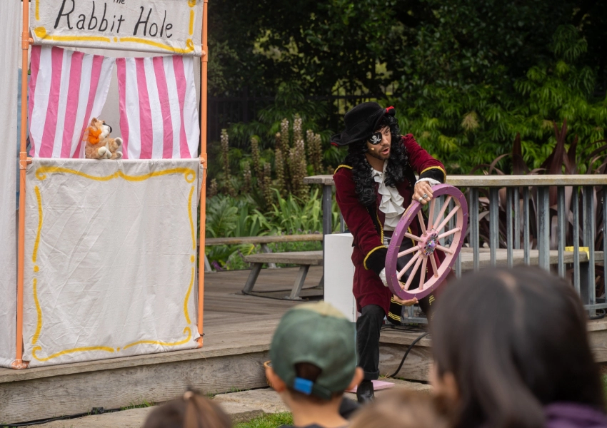 A performer in a pirate costume holds a large wheel on stage near a sign that reads "The Rabbit Hole," with an audience watching.