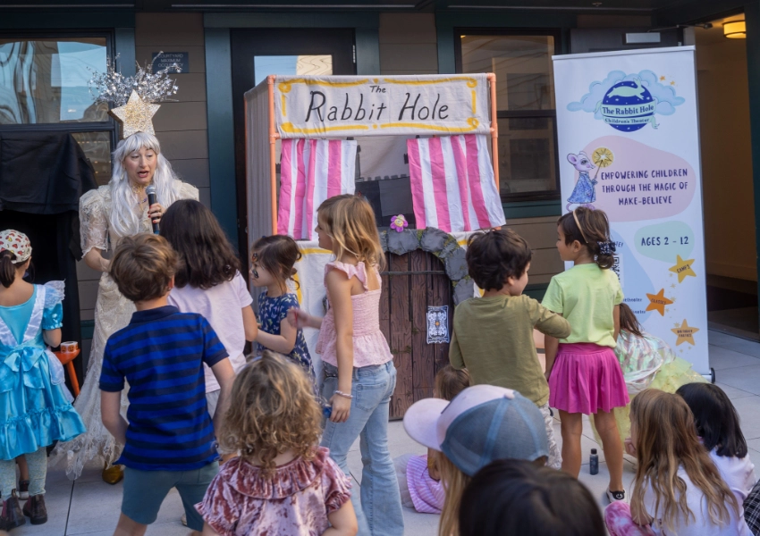 A performer dressed as a fairy entertains a group of children in front of a puppet theater setup labeled "The Rabbit Hole.