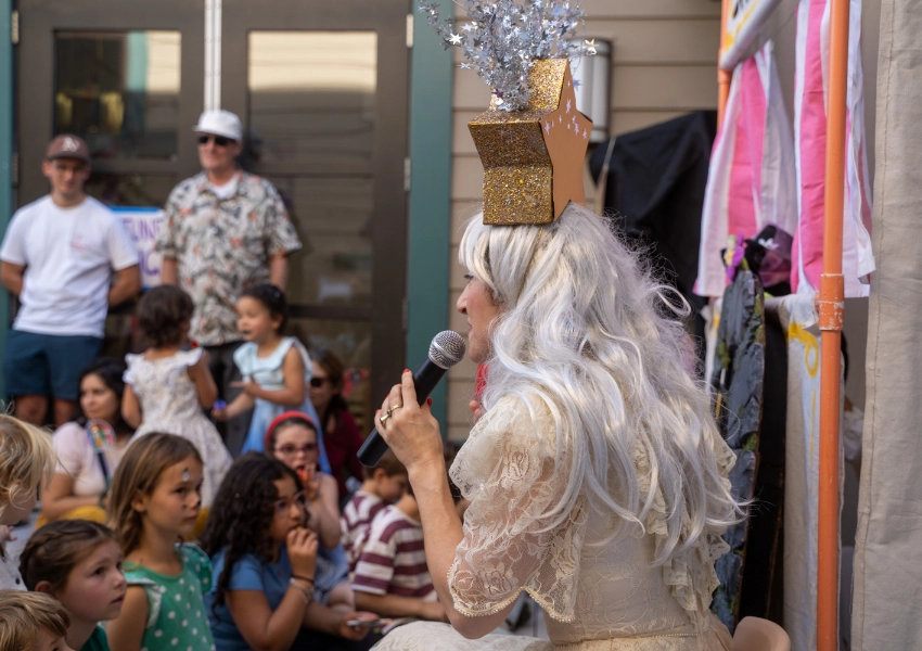 A performer in a shimmering outfit and white wig sings into a microphone onstage. Children sit and watch. Posters and audience members in casual attire are visible in the background.