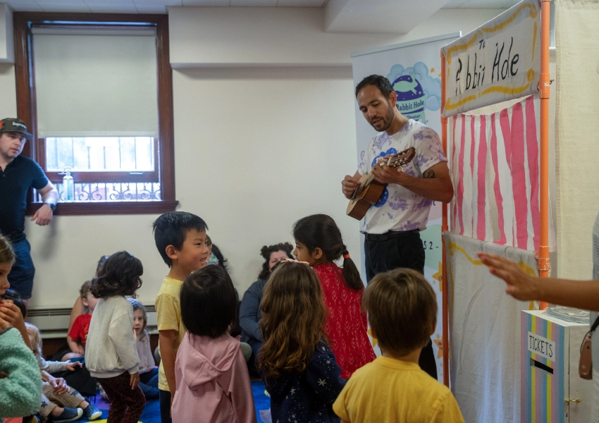 Man playing a ukulele in front of young children inside a room. A striped backdrop and a box labeled "Tickets" are visible.