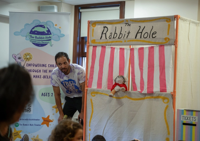 A man stands beside a puppet show booth labeled "The Rabbit Hole" with a puppet on display. A small audience is visible in the foreground.