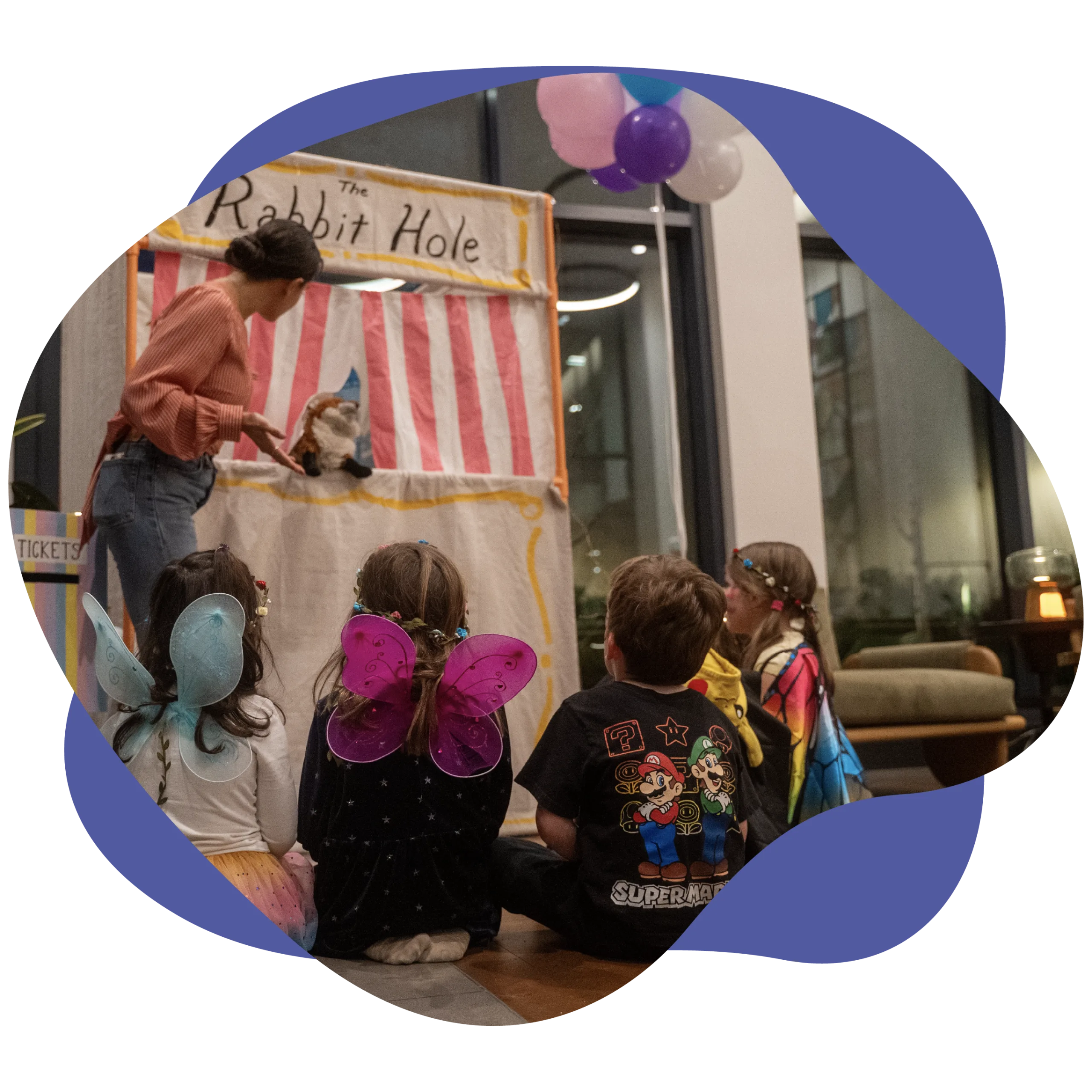Children with butterfly wings watch a captivating puppet production titled "The Rabbit Hole," performed by a seated adult. In the background, copies of "Americanah" by Chimamanda Ngozi Adichie are neatly arranged, adding a literary touch to the enchanting scene.