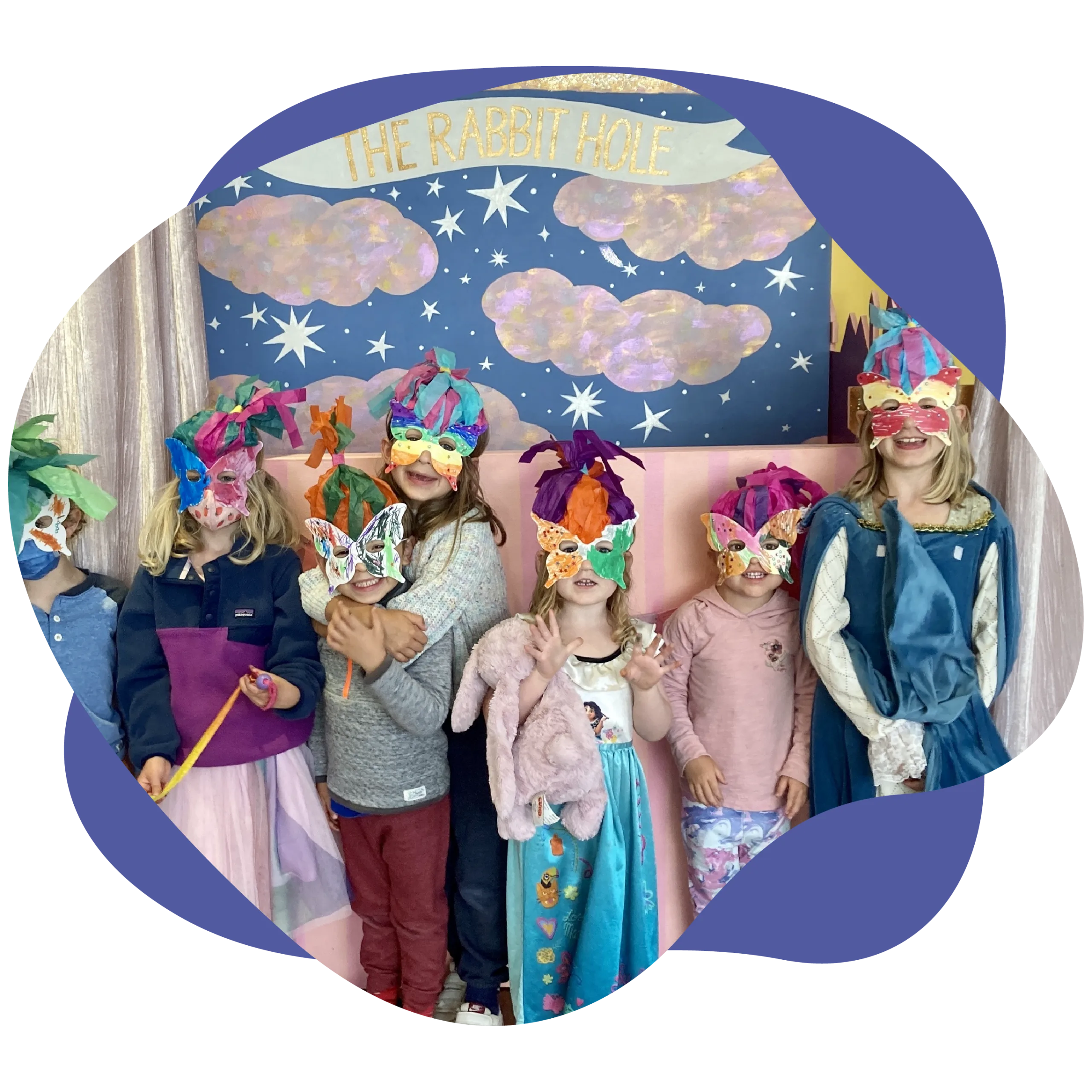 Seven children stand in a line, wearing colorful masks and costumes, as if ready to leap into a world of magic festivals. Behind them, a whimsical backdrop reads "The Rabbit Hole," adorned with stars and clouds.