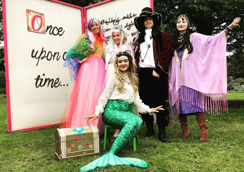 Five people in fairy tale costumes pose in front of a "Once upon a time..." backdrop on grass.