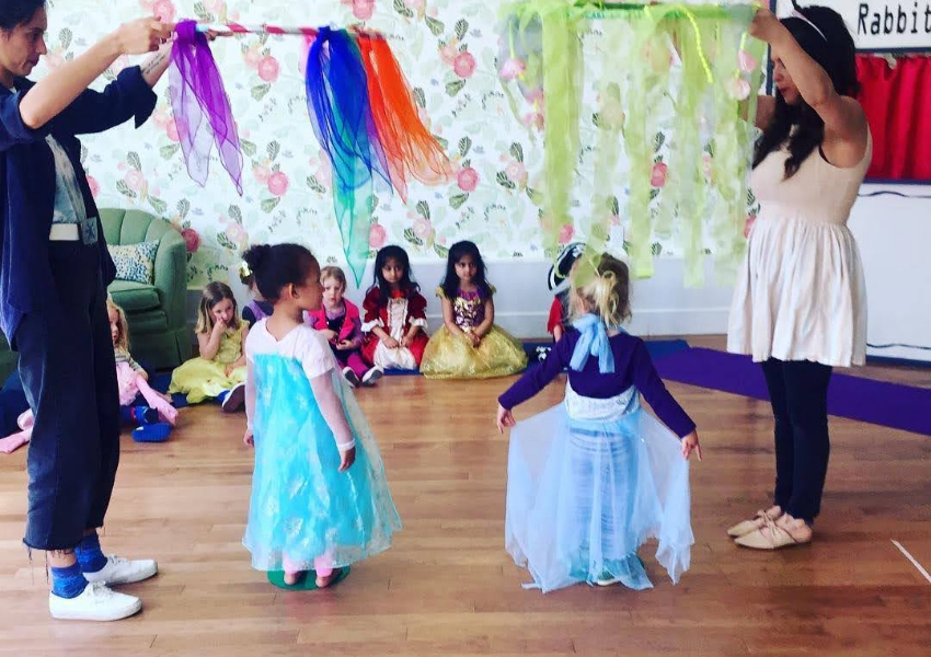 Children in costumes participate in an activity with colorful scarves, assisted by two adults, in a room with other seated children.