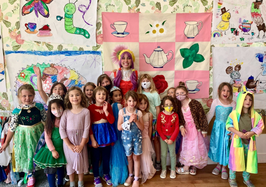 A group of children in colorful costumes pose for a photo with an adult in a bright outfit in front of walls covered with various drawings and paintings.