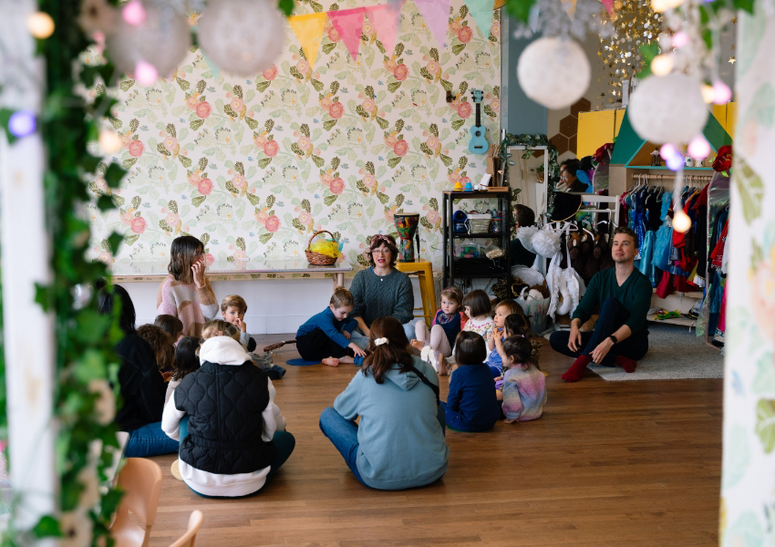 Children and adults sitting in a circle on a wooden floor in a playroom with floral wallpaper and colorful decorations.