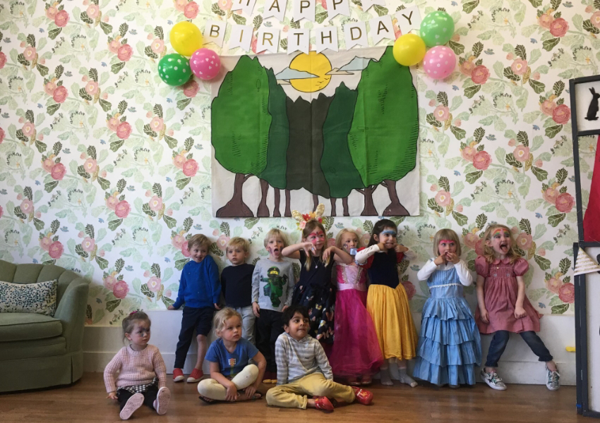 Children dressed in various costumes pose for a photo in front of a "Happy Birthday" banner, colorful balloons, and a wall decorated with a tree mural.