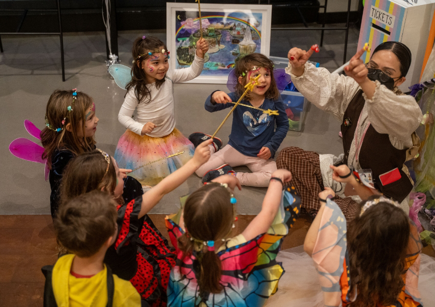 Children wearing costumes sit in a circle, playing a game. An adult gestures and interacts with them.