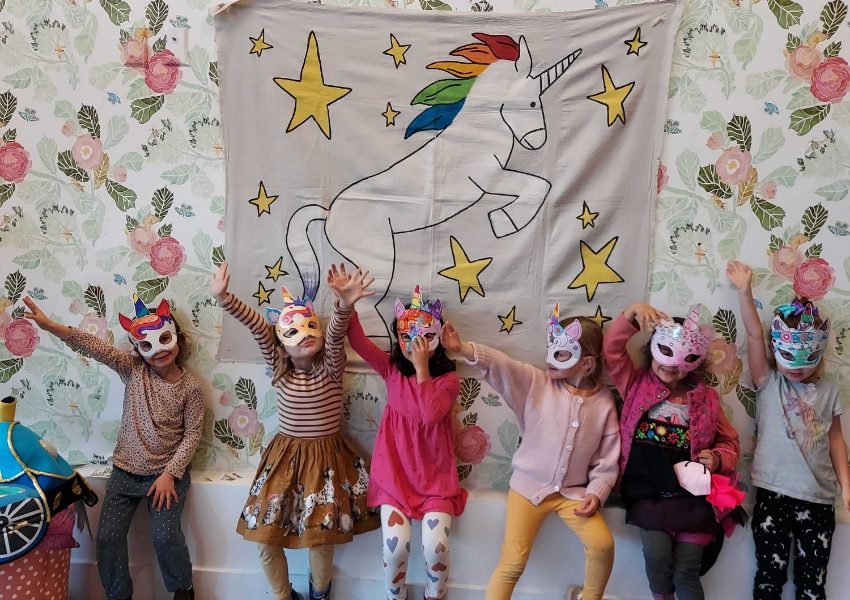 Six children wearing unicorn masks pose with arms raised in front of a unicorn tapestry on a floral-patterned wall.