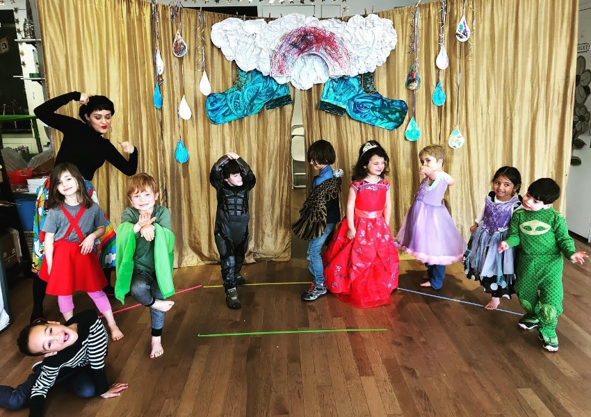 Children in costumes pose on a wooden floor in front of a gold curtain with cloud and drop decorations. An adult in black is standing to the left with one arm raised.
