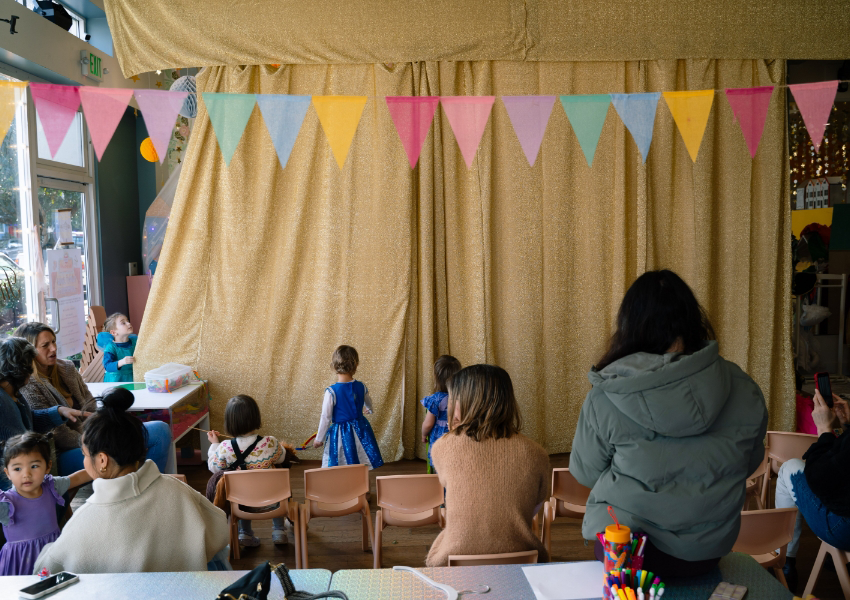 Children and adults in front of a gold-curtained stage with colorful bunting above, inside a room with chairs and a table.