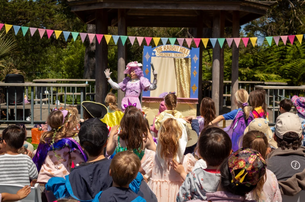 A performer in a pink costume entertains a group of children in costumes at an outdoor event called "The Rabbit Hole Children's Theatre." Colorful bunting decorates the stage.