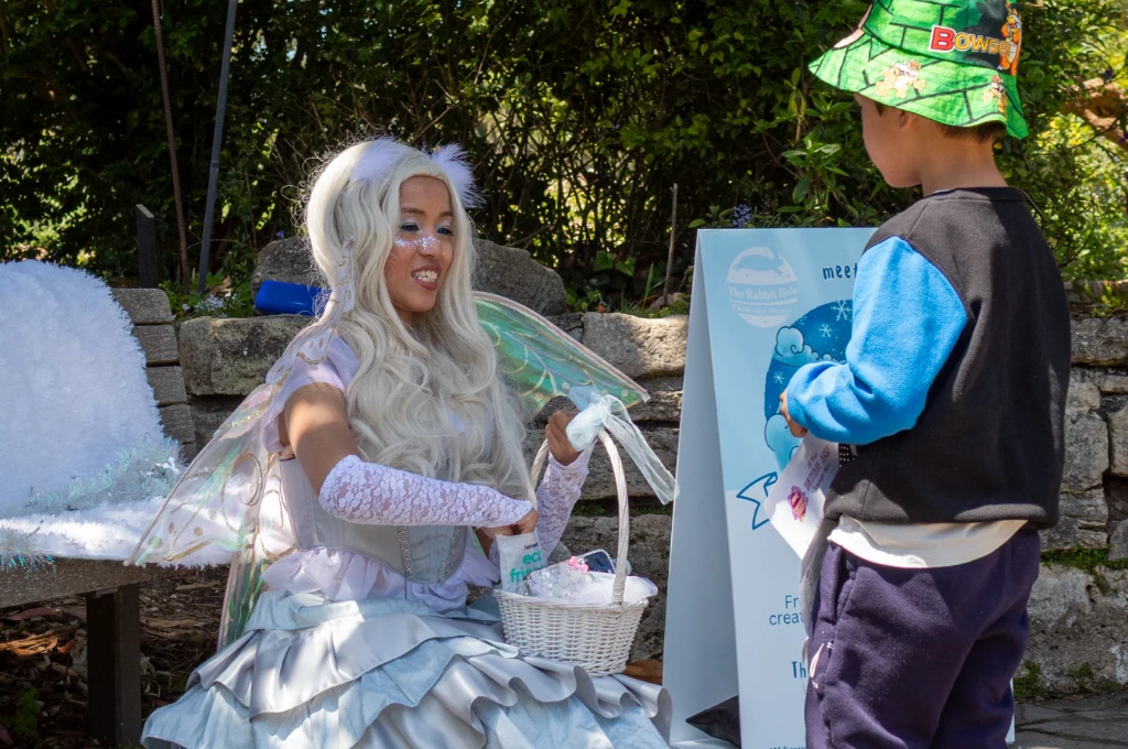 A person in a fairy costume with a white wig and wings hands something from a basket to a child wearing a green hat and blue shirt outdoors.