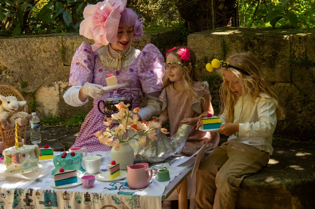 An adult in a pink costume and two children enjoy a tea party outdoors, holding colorful toy cake slices and cups at a decorated table.