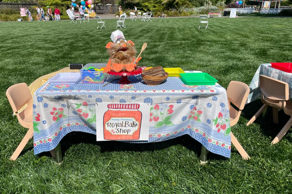A table with a “Royal Bake Shop” sign is set up outdoors, covered with a floral cloth, a puppet chef, baking props, and empty chairs around it.