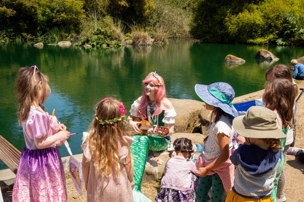 A performer dressed as a mermaid plays a ukulele for a group of children in costumes near a pond surrounded by greenery.