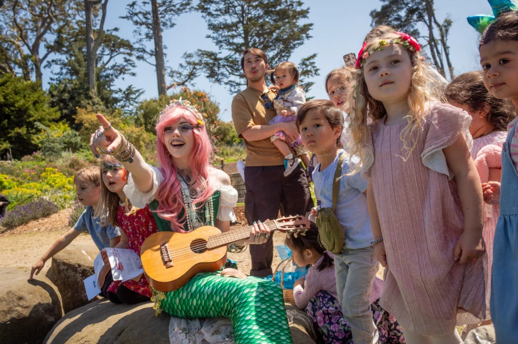 A woman in a mermaid costume with a ukulele sits on a rock, surrounded by children outdoors on a sunny day.
