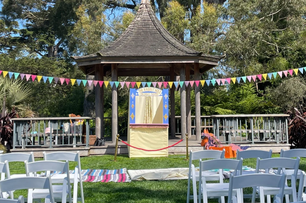 Outdoor puppet show setup with a small stage and puppet theater in front of a gazebo, colorful bunting overhead, and rows of white chairs for the audience.