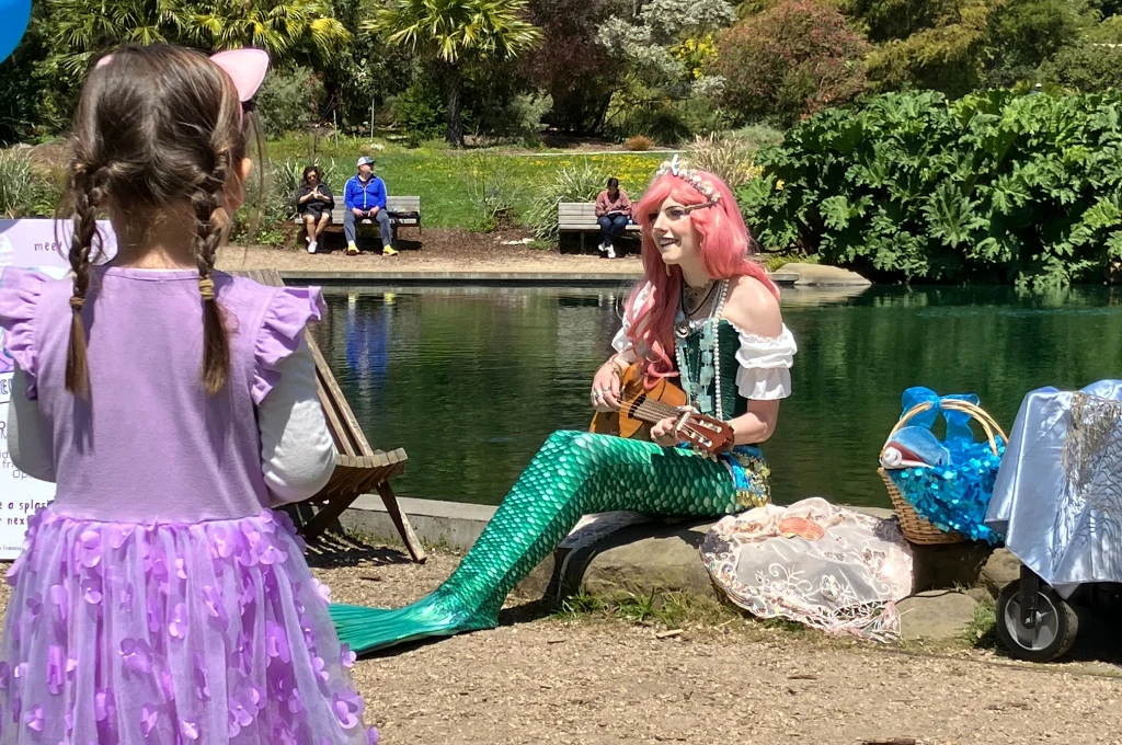 A woman dressed as a mermaid with a guitar sits by a pond, performing for a young girl in a purple dress and cat ears in a park setting.