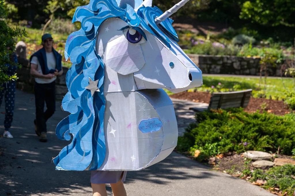 A person wearing a large, blue and white unicorn costume walks outdoors on a sunny day in a park setting.