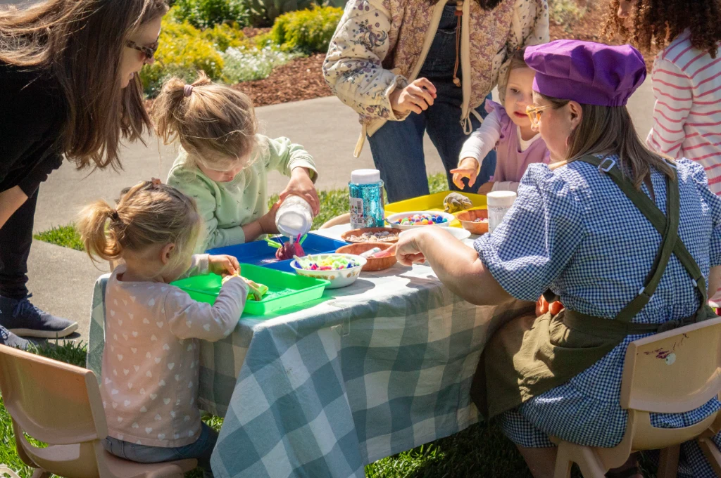 A group of children and two adults sit around a table outdoors, engaged in arts and crafts activities with colorful materials on trays.