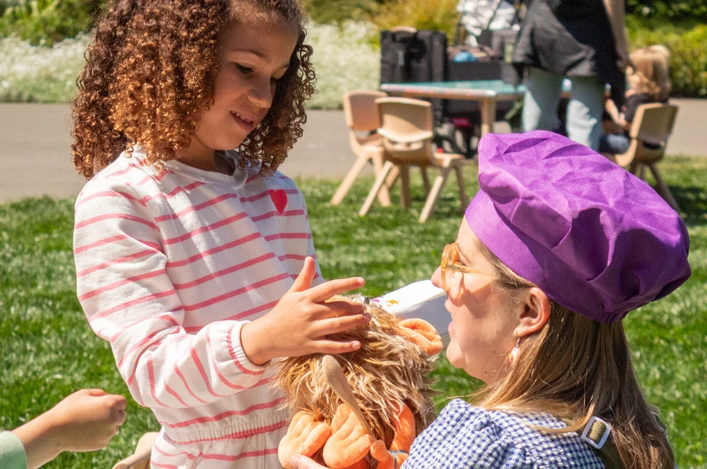 A young girl touches a stuffed animal held by a woman in a purple hat and sunglasses, outdoors on a sunny day.