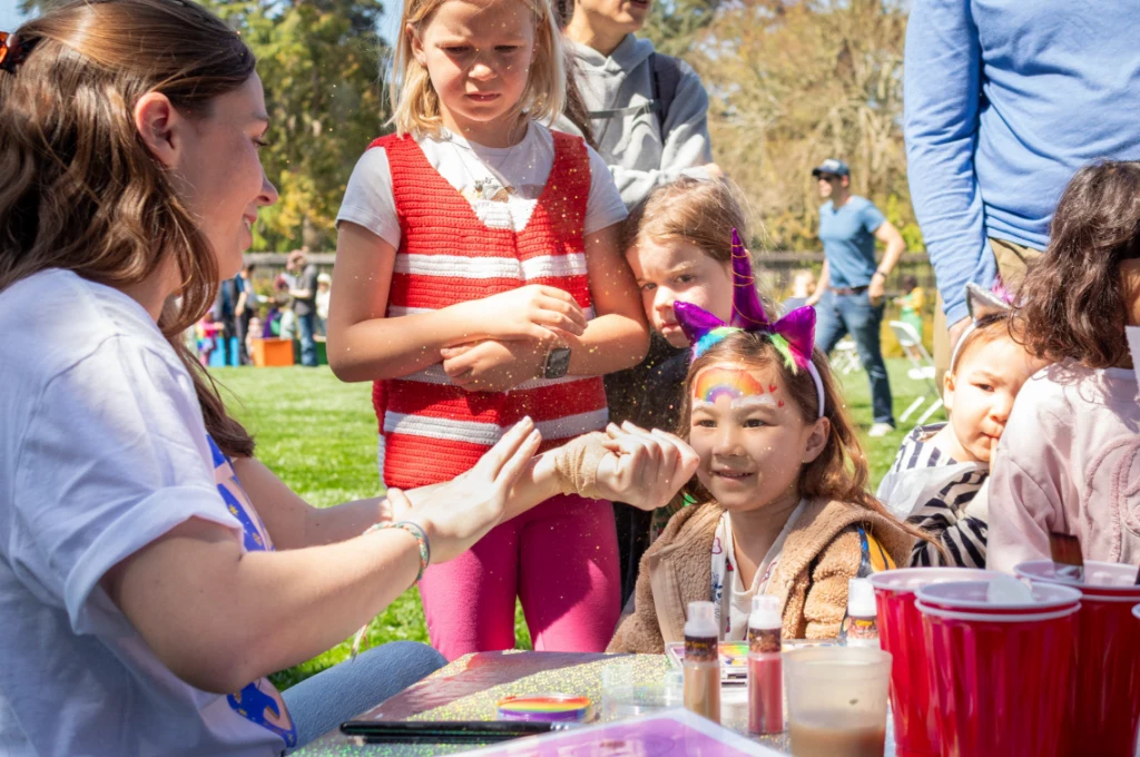 A woman sprinkles glitter on a smiling girl’s hand at an outdoor face painting booth, with other children waiting in line on a sunny day.
