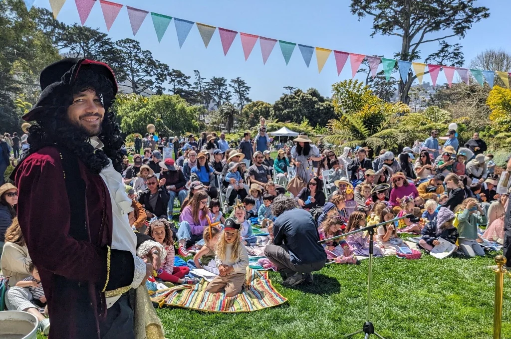 A man in a pirate costume stands in front of a large seated crowd outdoors on a sunny day, with colorful bunting hanging above.