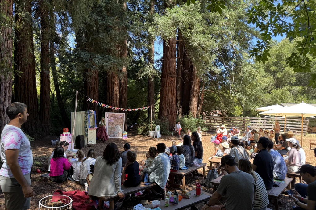 A group of people sit on benches outdoors, watching a puppet show in front of tall redwood trees. Colorful bunting hangs above the stage area.