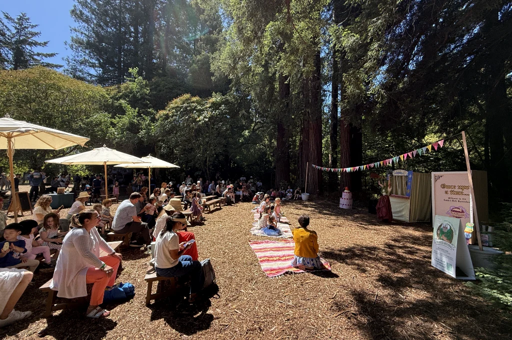 People sit on benches and blankets in a wooded outdoor area, watching a small stage with performers and colorful bunting under tall trees.