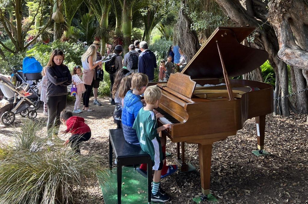 Children play a grand piano outdoors under trees while a group of people, including adults and children, gather nearby in a park-like setting.
