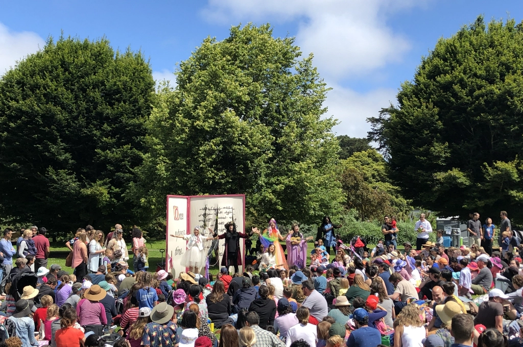 A large crowd sits outdoors watching a group of costumed performers on a stage set up in front of trees on a sunny day.