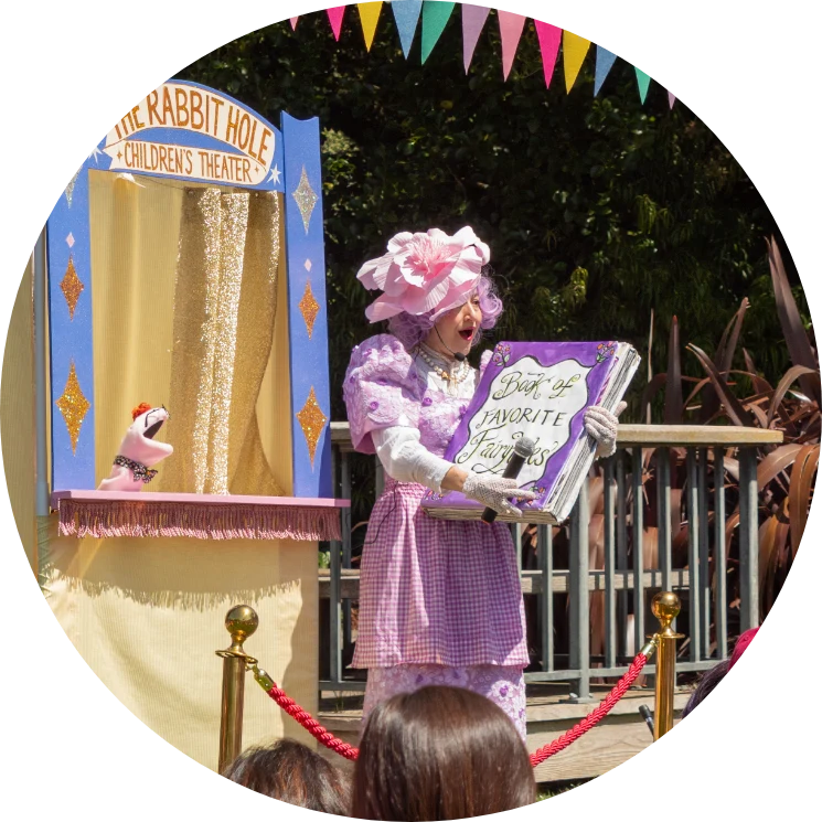 A performer in a pink costume holds a large book in front of a puppet theater labeled "The Rabbit Hole Children's Theater" during an outdoor event.