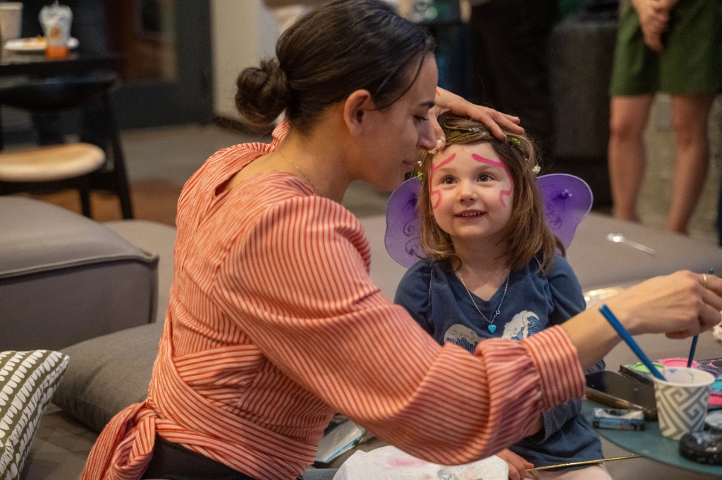An adult paints a young girl's face as she sits smiling, wearing purple butterfly wings and a headband, in a casual indoor setting.