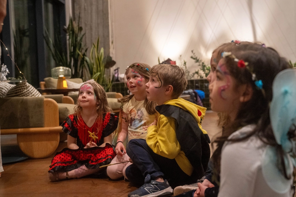 A group of young children in costumes sit on the floor indoors, attentively watching something out of view.