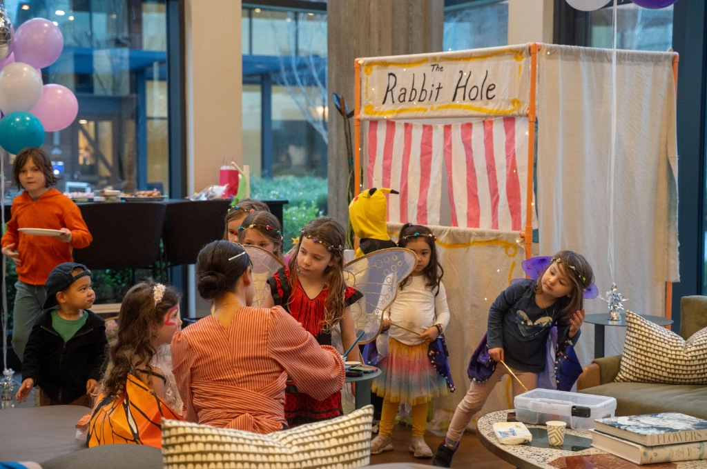 Children in costumes gather around a woman at a booth labeled "The Rabbit Hole" during an indoor event; balloons and decorations are visible in the background.