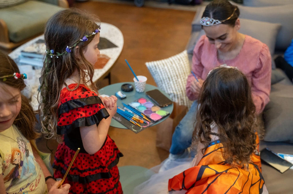 Three children in costume stand near a woman who is painting a girl’s face. Face paint supplies and brushes are on a small table beside them.