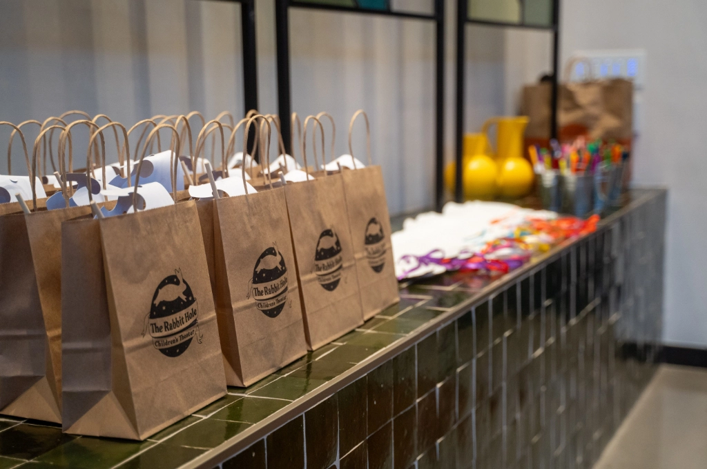 A row of brown paper gift bags with handles sits on a tiled counter, alongside colorful supplies and yellow vases in the background.