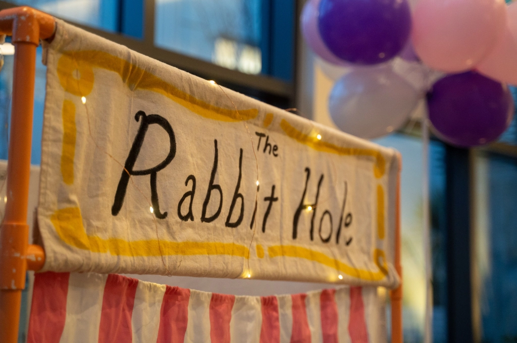 A sign reading "The Rabbit Hole" is displayed above red and white striped fabric, with string lights and purple and white balloons in the background.