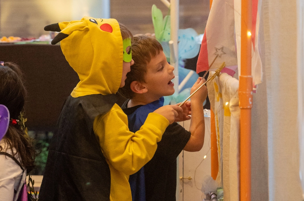 Two children in costumes, one dressed as Pikachu, stand closely together and interact with decorations hanging on a wall at a festive indoor event.