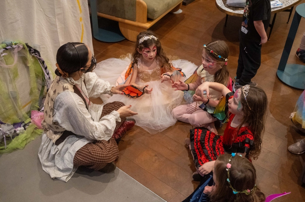 An adult in costume sits on the floor and gestures with their hands while four young children in fairy or princess costumes listen and watch attentively.